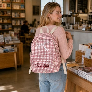 May include: A pink and white gingham patterned backpack featuring a white ribbon bow graphic on the upper section and the name Marina embroidered in dark pink cursive script on the front pocket, equipped with adjustable off-white shoulder straps.