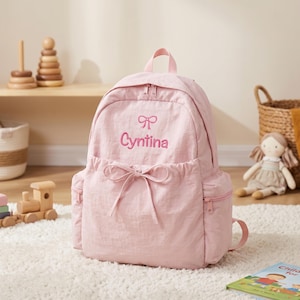 May include: A light pink backpack featuring a top handle, two side pockets with zips, and a front pocket with a drawstring tie. The front is embroidered with a small bow icon above the name Cyntina in a matching pink script font.