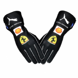 Peut inclure: Une paire de gants de course noirs avec des logos Puma blancs, le texte Richard Mille, un écusson Ferrari jaune et un logo Shell rouge et jaune sur le dos de chaque main. Les gants présentent des coutures blanches le long des doigts et poignets.