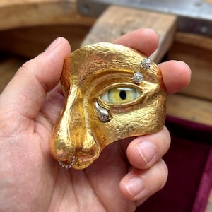 以下が含まれることがあります： 金色の彫刻された顔のペンダント。黄色い虹彩と銀色の涙を持つ開いた目があります。目の上には2つの小さなダイヤモンドアクセントがあり、鼻にはダイヤモンドの列があります。ペンダントはテクスチャ加工された表面を持っています。