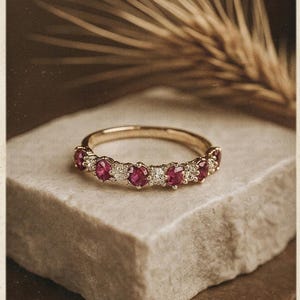 Puede incluir: Un anillo de oro con gemas alternas de rubí y diamante. El anillo está colocado sobre una piedra de color claro, con un fondo borroso de espigas de trigo. El anillo es de estilo media eternidad, con las gemas engastadas alrededor de la mitad superior de la banda.
