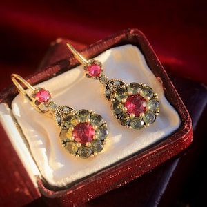 May include: A pair of gold drop earrings featuring a central round red gemstone surrounded by a halo of smaller olive-green stones. Each earring has a smaller red stone at the top and a lever-back closure, presented in a dark red jewelry box.