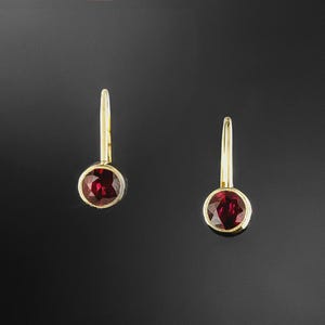 Peut inclure: Une paire de boucles d'oreilles pendantes en or avec des pierres précieuses rondes et facettées d'un rouge profond, serties dans une monture en chaton. Chaque boucle possède un crochet en fil d'or fin et incurvé, créant un design épuré.