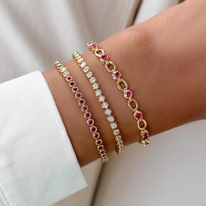 Könnte beinhalten: Drei zusammen getragene Goldarmbänder. Das erste ist ein Tennisarmband mit abwechselnd roten und klaren Steinen. Das zweite ist ein Tennisarmband mit klaren Steinen. Das dritte ist ein Goldkettenarmband mit ovalen Gliedern, die mit roten Steinen besetzt sind.