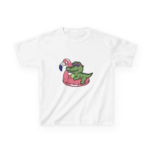 Könnte beinhalten: Weißes T-Shirt mit einem Cartoon-Alligator mit Sonnenbrille, der auf einem rosa Flamingo-Schwimmring schwimmt. Der Alligator ist grün, und der Schwimmring hat einen rosa Körper mit einem blauen und schwarzen Schnabel. Das Design ist zentriert.