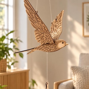 May include: A hanging wooden bird ornament featuring intricate laser-cut feather details and a natural brown finish. The bird is suspended by thin white strings and includes a small, matching wooden weight attached to the underside for balance.