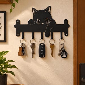 Puede incluir: Un soporte para llaves de pared de metal negro con la silueta de un gato asomándose por encima de la barra superior con una pata colgando. El estante incluye cinco ganchos, cada uno con un juego de llaves, incluyendo un mando de coche.