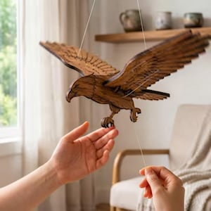 Pode incluir: Um autômato de águia em madeira castanho-escura com asas e garras articuladas, suspenso por fios brancos finos. A ave apresenta padrões de penas detalhados cortados a laser e foi concebida para bater as asas ao puxar o fio de controlo.