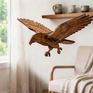 May include: A hanging wooden eagle sculpture with outstretched wings, crafted from layered brown wood pieces. The bird is depicted in a mid-flight pose with its talons extended downward, showcasing detailed feather patterns and a natural wood grain finish.