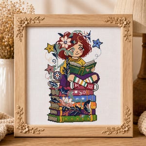 May include: A cross-stitch embroidery in a light wood frame with floral corner carvings. The design features a person with reddish-brown hair reading a green book while sitting on a stack of colorful books, surrounded by stars and flowers.