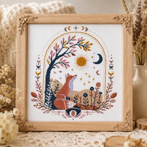 May include: A square cross-stitch embroidery in a light wood frame with floral corner carvings. The design features a fox sitting under a tree with pink leaves, a yellow sun, a blue crescent moon, and stars, all enclosed within an arched border.