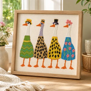 May include: A framed cross-stitch embroidery featuring four stylized geese standing in a row. Each goose wears a different hat and a uniquely patterned outfit, including green waves, leopard print, black spots, and blue with pink flowers.