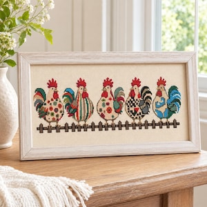 Puede incluir: Obra de arte en punto de cruz enmarcada con seis gallos coloridos sobre una valla marrón. Cada gallo tiene un patrón único de rojo, verde, azul y blanco. El marco es de color claro y neutro.