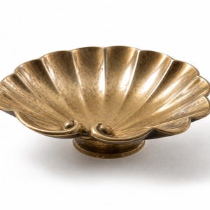 Könnte beinhalten: Eine dekorative, muschelförmige Schale aus Messing im antiken Stil. Die Schale hat einen gewellten Rand und steht auf einem kleinen, runden Sockel. Das Innere hat eine strukturierte Oberfläche und die Farbe ist goldfarben.