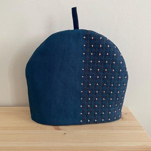 以下が含まれることがあります： 濃い青色の布で作られたドーム型のティーコージー。左側は無地で、右側には白い糸で刺し子風の幾何学模様が刺繍されています。上部中央には、小さな濃い青色の布製のループが付いています。