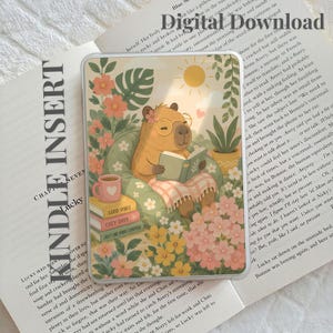 Puede incluir: Una funda protectora transparente para lector electrónico Kindle con una ilustración impresa de un capibara con gafas leyendo un libro en un sillón verde, rodeado de flores y una pila de libros con las etiquetas Good Vibes, Cozy Days y Just One More Chapter.