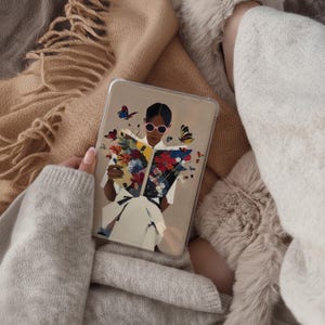 Könnte beinhalten: Transparente Tablet-Hülle mit einer Illustration einer Person, die ein Buch mit bunten Blumen und Schmetterlingen liest. Die Person trägt eine rosa Sonnenbrille. Die Hülle wird von einer Person gehalten, die einen grauen Pullover trägt und auf einer beigen Decke ruht.