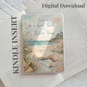 May include: A clear Kindle insert featuring a watercolor illustration of an open book on a sandy beach with the text Lost in a Good Book and a Better View. The scene includes coastal flowers, a seashell, a sand dollar, and a distant house.