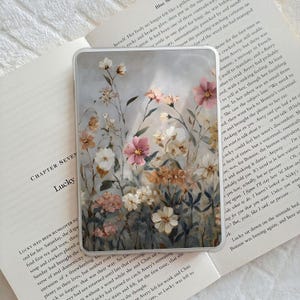 May include: Transparent phone case with a floral pattern in pink, white, and peach hues. The case is placed on an open book, with the word "Lucky" visible.