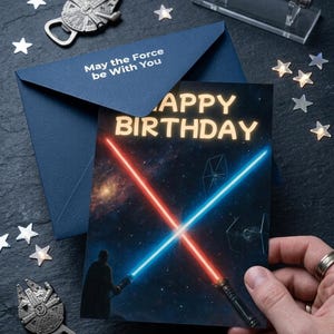 Peut inclure: Une carte d'anniversaire avec l'inscription dorée "HAPPY BIRTHDAY", représentant des sabres laser rouges et bleus croisés sur fond spatial. Une enveloppe bleue avec "May the Force be With You" est à côté de la carte. Des confettis en forme d'étoiles et un ouvre-bouteille Faucon Millenium sont également présents.