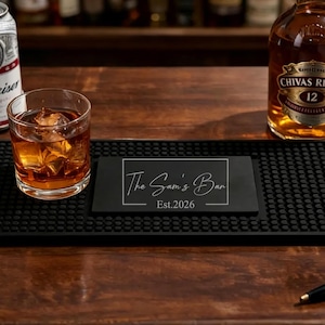 May include: A rectangular black rubber bar mat featuring a textured surface with raised nubs and a central smooth panel. The panel displays the white text "The Sam's Bar" enclosed in a rectangular border, with "Est.2026" printed underneath the name.