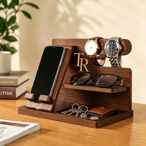 Op de afbeelding: Een bureau-organizer van donker hout met een smartphonehouder, een horizontale plank voor een bril en een basisplateau voor sleutels en kleine voorwerpen. De unit toont de goudkleurige letters TR, met twee polshorloges aan de bovenkant.