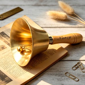 May include: A brass hand bell with a polished gold-tone finish and a light-coloured wooden handle. The handle features the name Nancy engraved in a dark script font. A small metal clapper hangs inside the bell, which is resting on a book.