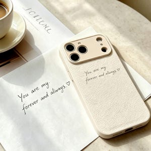 May include: A cream-coloured smartphone case with a textured surface and a camera cutout, featuring the printed text 'You are my forever and always.' with a small heart symbol. The case is placed on a white paper with the same printed text.