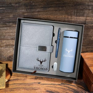 Peut inclure: Un coffret cadeau dans une boîte grise, comprenant un carnet gris avec un fermoir argenté, un stylo bleu et un thermos bleu clair. Le carnet et le thermos portent un logo de tête de cerf et le nom "THOMAS".