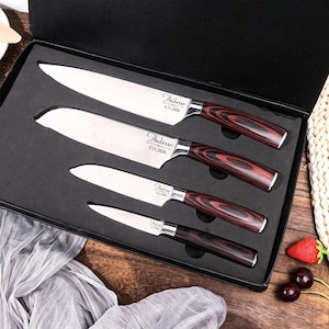 Peut inclure: Un ensemble de quatre couteaux de cuisine en acier inoxydable avec des manches en bois veiné rouge foncé et noir, présentés dans une boîte noire doublée de mousse. Chaque lame est gravée du nom Andrew et de la date 5.11.2026 en écriture cursive.