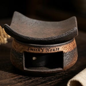 May include: A dark brown ceramic wax warmer with a textured, rustic finish and a curved top dish. The front features a rectangular opening and the text Smith's House engraved in a dark font. The base is rounded with a gradient of brown tones.