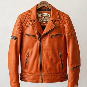 Könnte beinhalten: Eine orangefarbene Motorradjacke aus Leder mit gesteppten Schulterpartien, einem zentralen Reißverschluss, mehreren Reißverschlusstaschen und verstellbaren Seitenschnallen. Das Innenetikett zeigt den Text ADVENTURE AWAITS Est. 2023 und ein Aufnäher am Ärmel trägt die Aufschrift WILDHEART.