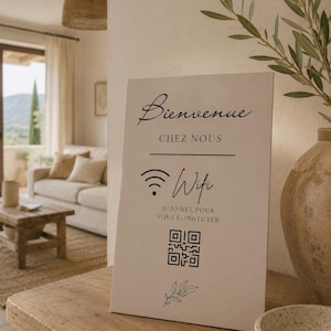 Op de afbeelding: Een rechthoekig bord met de woorden "Bienvenue Chez Nous" in sierlijk schrift, samen met een Wi-Fi-symbool en een QR-code. Het bord staat op een houten oppervlak, met een neutraal getint interieur op de achtergrond.