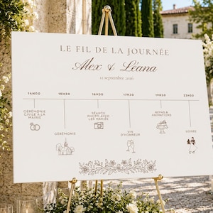Peut inclure: Un panneau de planning de mariage blanc sur un chevalet doré avec le texte LE FIL DE LA JOURNÉE, Alex & Léana, et 12 septembre 2026. Le panneau affiche un programme chronologique avec des icônes pour la cérémonie, les photos et la soirée.
