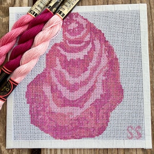 May include: A needlepoint canvas featuring a pink rose design, with varying shades of pink. Three skeins of embroidery floss in coordinating pink hues are positioned above the canvas. The canvas is on a white background.
