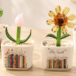 May include: Four glass flower figurines in square white ceramic pots. Each pot features a colorful illustration of a row of books and the text "Mrs. Smith". The flowers include a purple bell, a pink tulip, a yellow daisy, and white lily-of-the-valley.
