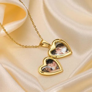 Puede incluir: Un collar con colgante en forma de corazón de tono dorado con una cadena delicada. El colgante está abierto, revelando dos compartimentos interiores separados, cada uno con una pequeña fotografía de una pareja abrazada dentro del marco metálico.