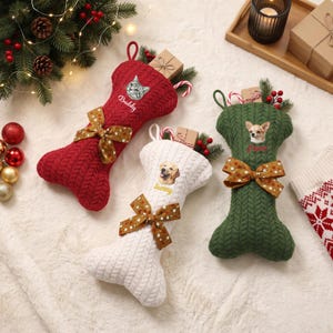 Puede incluir: Tres calcetines navideños con forma de hueso en tejido de punto trenzado rojo, blanco y verde. Cada calcetín presenta un retrato de mascota, un nombre bordado en cursiva y un lazo marrón con lunares. Los nombres Buddy, Sunny y Luna son visibles.