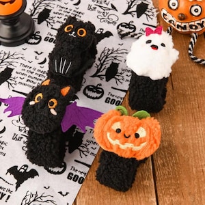 Op de afbeelding: Vier pluche Halloween-slap-armbanden met een zwarte kat met oranje ogen, een zwarte vleermuis met paarse vleugels, een witte geest met een roze strik en een oranje pompoen. De armbanden liggen op een witte stof met zwarte Halloween-tekst en illustraties.