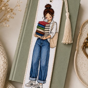 Puede incluir: Un marcapáginas rectangular de punto de cruz que muestra a una persona con un moño castaño, vistiendo una camisa blanca y vaqueros azules, sosteniendo una pila de cinco libros coloridos. El marcapáginas tiene un borde de tela crema y una borla a juego.