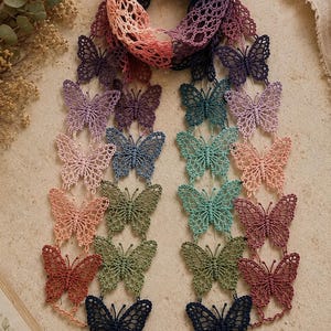 Peut inclure: Une écharpe au crochet composée d'une série de motifs de papillons interconnectés dans un dégradé de couleurs incluant pêche, lavande, sarcelle, vert olive et bleu foncé. L'écharpe présente une structure ajourée avec des détails en fil.