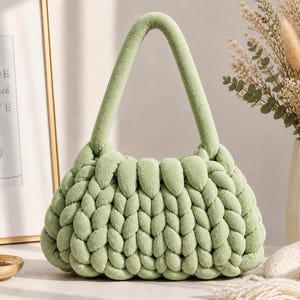 May include: A pale green, knitted handbag with a rounded shape and a soft, textured appearance. The bag has a matching handle and a woven design. The bag is displayed near a framed print that says "MADE with LOVE".