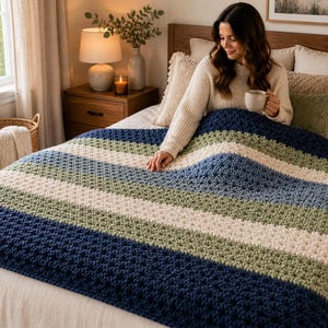Könnte beinhalten: Eine grob gehäkelte Decke mit horizontalen Streifen in Marineblau, Salbeigrün und Creme. Die Decke hat eine texturierte, dicke Garnstruktur mit einem sich wiederholenden Stichmuster und bedeckt die Bettoberfläche mit einem weichen Aussehen.