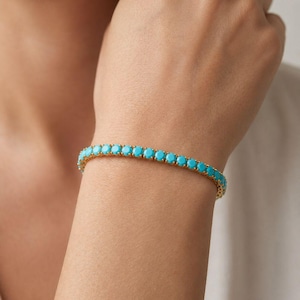Könnte beinhalten: Ein goldfarbenes Tennisarmband mit einer durchgehenden Reihe runder, leuchtend türkisfarbener Steine, die in einzelnen Metallfassungen sitzen. Das Armband umschließt das Handgelenk und zeigt ein einheitliches Design mit blau-grünen Edelsteinen.