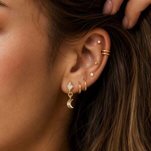 May include: A collection of gold-toned earrings worn in a single ear, featuring a dangling moon and star charm with opal-like stones, two small plain hoop earrings, a tiny star stud, a small ball stud, a double-band ear cuff, and a small crystal stud.