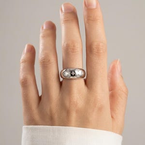 Puede incluir: Un anillo de banda en tono plateado pulido con un perfil ancho y redondeado. La superficie del anillo está decorada con múltiples motivos de estrellas de cuatro puntas grabados, cada uno centrado con una pequeña piedra incrustada brillante.