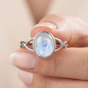 Peut inclure: Une bague argentée ornée d'une pierre de lune ovale centrale présentant un éclat bleu translucide et irisé. L'anneau présente un design divisé qui se rétrécit en un détail métallique torsadé de chaque côté du sertissage lisse et poli.