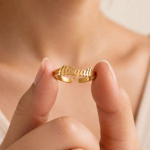 May include: A gold-toned metal ring featuring the name Abigail in a cursive script font. The ring has an open-ended band design, allowing for adjustment. The surface of the metal has a polished, reflective finish that catches the light.