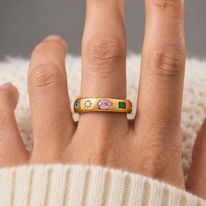 May include: A gold-toned band ring featuring a series of embedded gemstones and motifs. The design includes a small blue round stone, a star-shaped engraving with a central clear stone, a pink pear-shaped stone, and a square green stone.