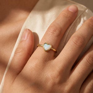 May include: A gold ring featuring a heart-shaped opal stone with iridescent white and blue tones. The band is thin and polished, holding the heart-shaped setting securely in place. The stone displays a smooth surface with subtle color play.